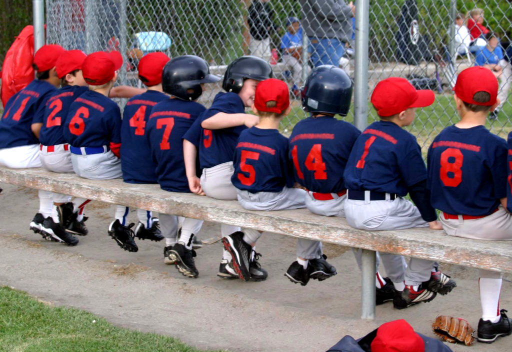 Young baseball players in red and blue uniforms sitting on a bench in a dugout.